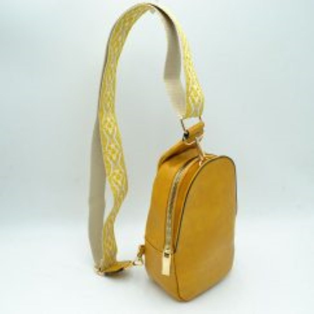 Mustard This compact sling backpack features a smooth textured finish and a contoured, teardrop-shaped silhouette designed for casual everyday use. It includes a primary storage compartment that secures with a prominent gold-tone zipper and a flat pull tab for easy access to essentials. A distinctive, wide shoulder strap decorated with an intricate geometric pattern connects via gold-tone metal clips to various anchor loops, allowing for adjustable and versatile cross-body or over-the-shoulder wear.