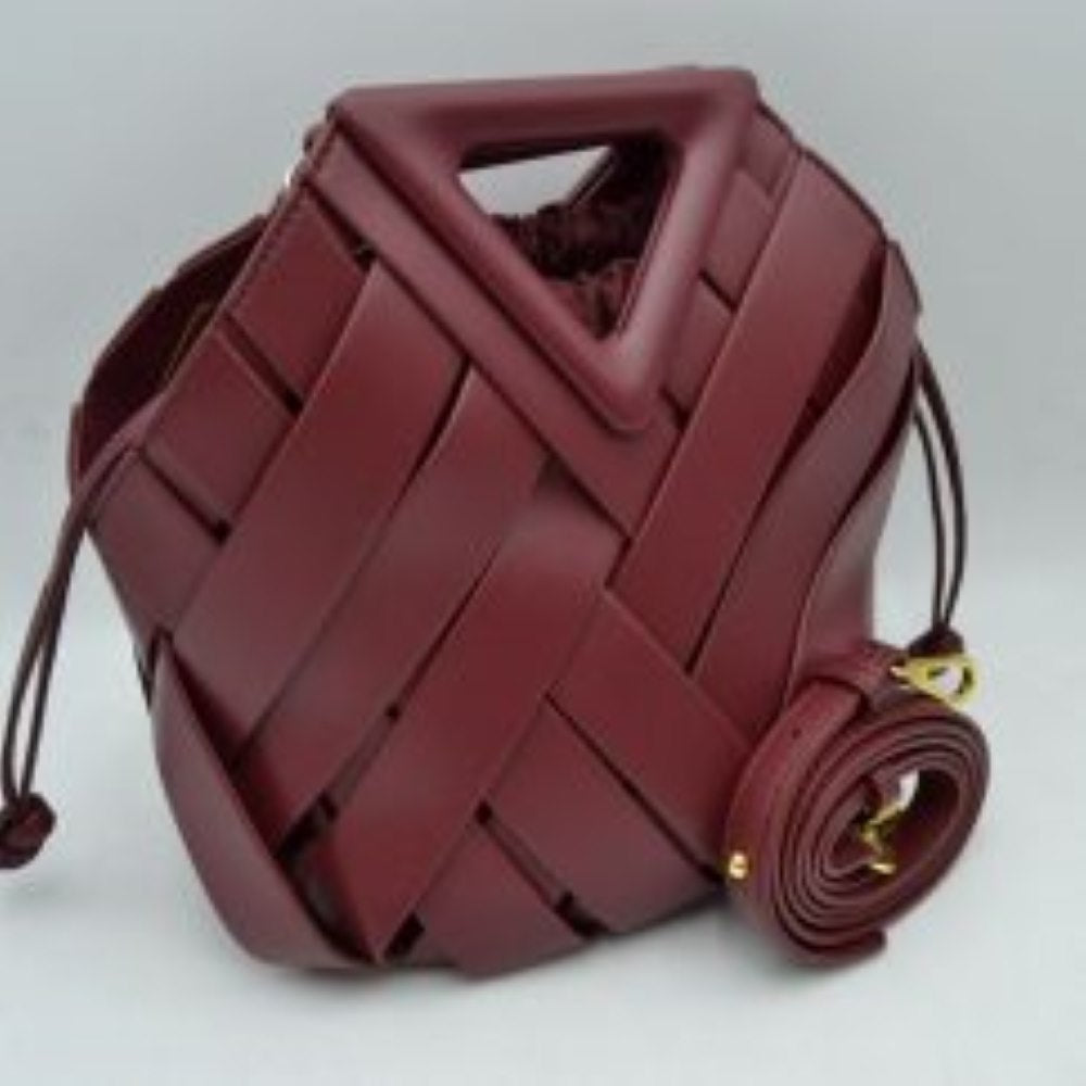 Wine This structural bucket bag features a distinctive geometric design composed of overlapping wide woven straps that create an eye-catching pattern, making it a unique statement piece for everyday use. A rigid triangular cutout shape forms an integrated top handle, while an additional long, flat shoulder strap extends from the upper frame for comfortable over-the-shoulder carrying.