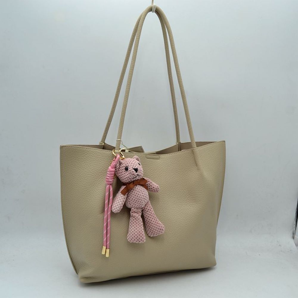 Beige his classic tote bag features a subtly pebbled exterior texture with a relaxed yet structured silhouette that is ideal for everyday use. Designed with a clean, open top for convenient access to the spacious main storage compartment, the bag is equipped with dual long, slim shoulder handles securely anchored to the interior panels. A decorative plush bear charm with a tiny bow sits on the front