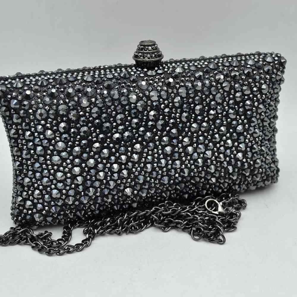 Black This elegant box clutch bag features an all-over array of faceted, shimmering rhinestone embellishments that fully cover the exterior for a textured, light-catching look. Designed for evening or special occasion use, the structured frame includes a secure top clasp closure with a tiered, decorative knob that easily snaps open to access the main storage compartment. A matching, detachable metallic link chain strap with a secure lobster clasp is included