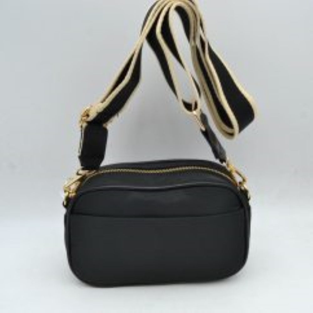 Black This compact camera-style crossbody bag features a smooth, minimal exterior and a rounded rectangular silhouette perfect for casual everyday use. It includes multiple functional storage compartments, consisting of a spacious main zippered section with a prominent gold-tone zipper and a convenient slip pocket across the front panel. A wide, adjustable fabric shoulder strap with contrasting light-colored borders attaches securely to gold-tone side loops