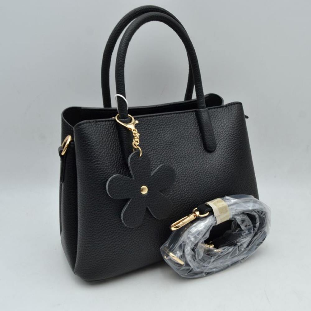 Black Add a touch of timeless elegance to your daily rotation with this beautifully structured satchel handbag. Crafted with a premium pebble-textured finish that balances durability with a polished look, this bag features a classic flared silhouette that sits securely on a flat base. The front handle is adorned with a charming, removable matching flower keychain dangle attached via a gleaming metallic clasp, adding a subtle playful note to a professional design.