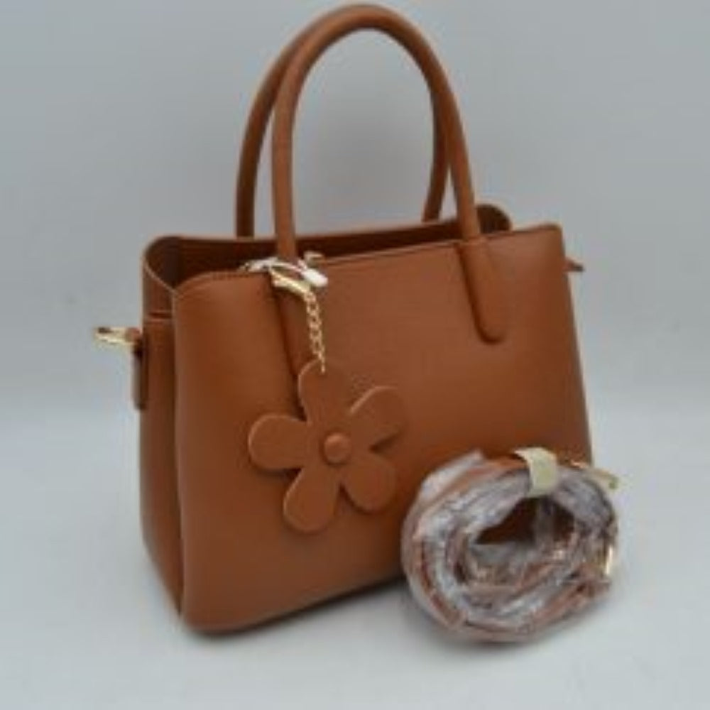 Brown Add a touch of timeless elegance to your daily rotation with this beautifully structured satchel handbag. Crafted with a premium pebble-textured finish that balances durability with a polished look, this bag features a classic flared silhouette that sits securely on a flat base. The front handle is adorned with a charming, removable matching flower keychain dangle attached via a gleaming metallic clasp, adding a subtle playful note to a professional design.