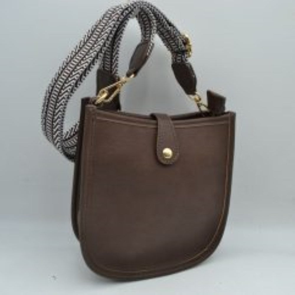 Coffee This stylish crossbody bag presents a clean, minimalist design with a smooth exterior finish and a distinct curved, U-shaped silhouette perfect for everyday use. It features an open top main compartment equipped with a small secure tab closure that fastens at the front with a metallic snap button. Adding a striking contrast, the bag is paired with a thick, adjustable fabric strap showcasing a bold geometric pattern