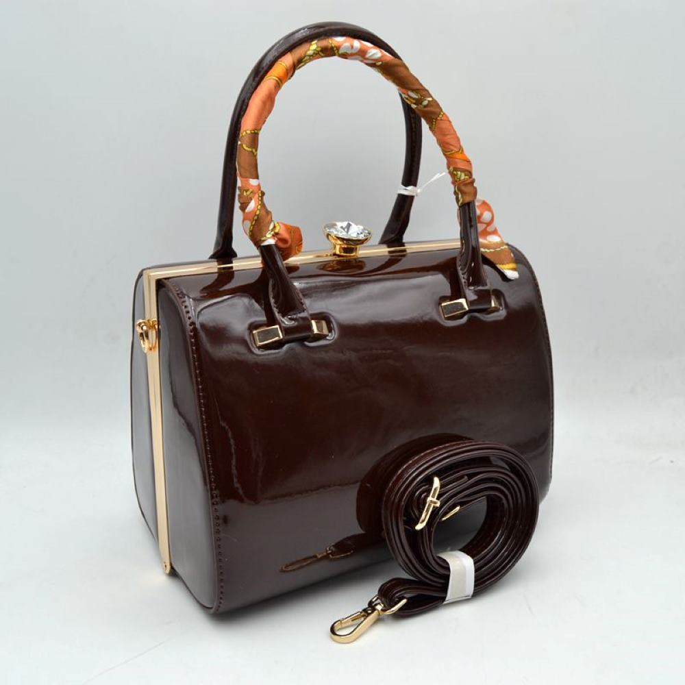 Coffee This structured handbag features a glossy finish and a rigid silhouette designed for everyday use. It is equipped with dual top handles, featuring a patterned decorative wrap on the front handle, and a decorative ornamental clasp closure at the top. The main storage compartment is accessed via a rigid frame opening, and the item includes a separate adjustable, detachable shoulder strap with clip attachments that connect to the side hardware.