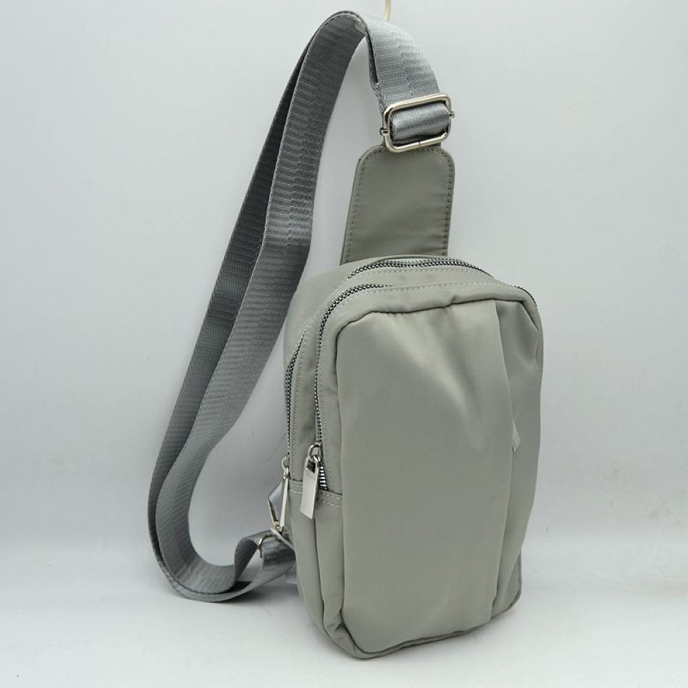 Gray This functional sling bag features a smooth, clean exterior with a structured vertical silhouette designed for active everyday use. It offers multiple organized storage compartments, including a primary back section and a secondary front pocket, both accessed via parallel top-curved zipper tracks with sleek metallic pulls. The bag is equipped with a wide, woven fabric shoulder strap attached to a integrated top panel and secured with an adjustable metallic buckle to easily customize the carrying length