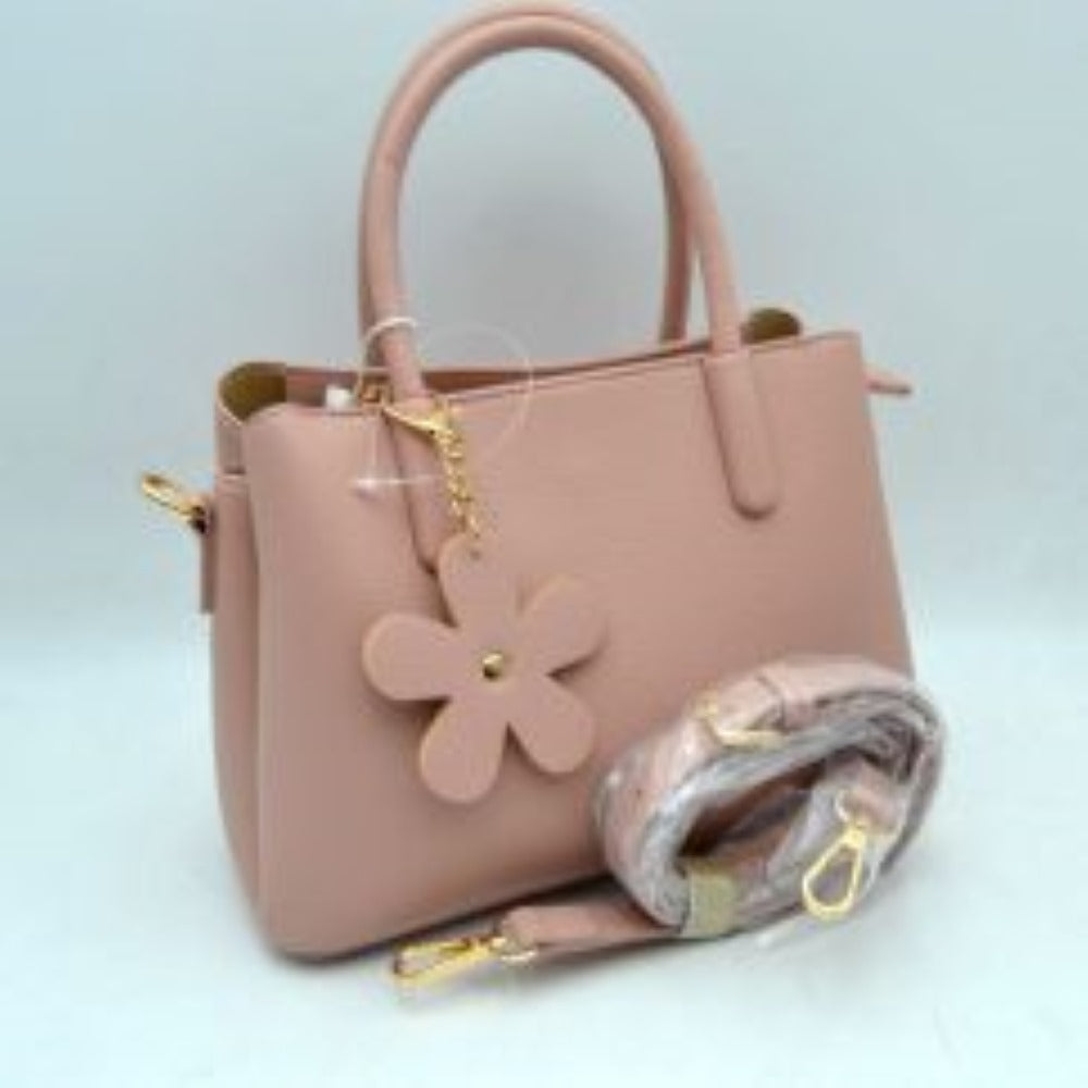 Pink Add a touch of timeless elegance to your daily rotation with this beautifully structured satchel handbag. Crafted with a premium pebble-textured finish that balances durability with a polished look, this bag features a classic flared silhouette that sits securely on a flat base. The front handle is adorned with a charming, removable matching flower keychain dangle attached via a gleaming metallic clasp, adding a subtle playful note to a professional design.