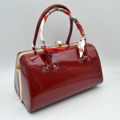 Red This structural handbag showcases a sleek barrel silhouette crafted from glossy patent leather in a rich burgundy red tone, offering a sophisticated aesthetic for daily or evening use. It features dual rounded top handles, with the front handle elegantly wrapped in a colorful patterned silk-like scarf accent tied neatly at the base. A rigid metallic gold-tone frame with a decorative top clasp opens wide to a secure main storage compartment, while side rings allow for the addition of a shoulder strap.