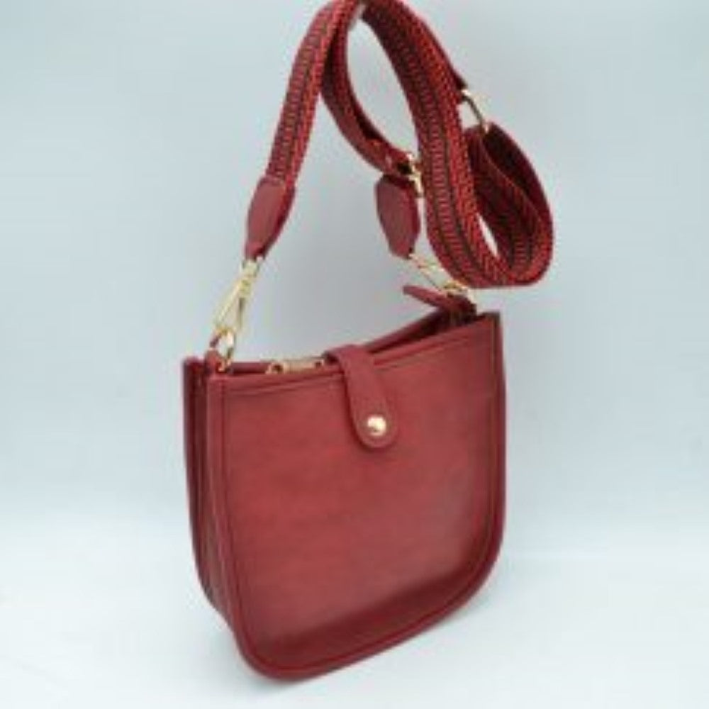 Red This stylish crossbody bag presents a clean, minimalist design with a smooth exterior finish and a distinct curved, U-shaped silhouette perfect for everyday use. It features an open top main compartment equipped with a small secure tab closure that fastens at the front with a metallic snap button. Adding a striking contrast, the bag is paired with a thick, adjustable fabric strap showcasing a bold geometric pattern