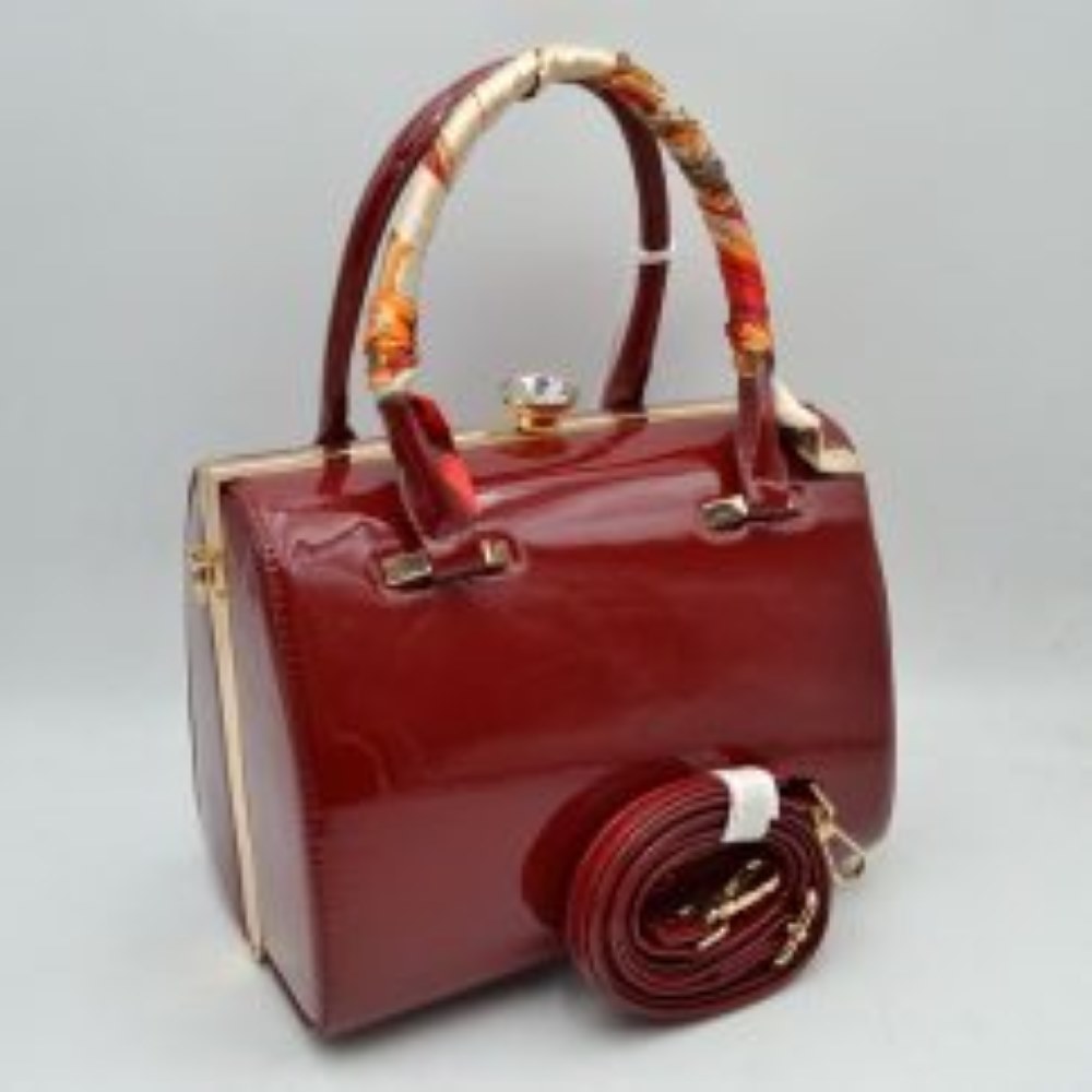 Red This structured handbag features a glossy finish and a rigid silhouette designed for everyday use. It is equipped with dual top handles, featuring a patterned decorative wrap on the front handle, and a decorative ornamental clasp closure at the top. The main storage compartment is accessed via a rigid frame opening, and the item includes a separate adjustable, detachable shoulder strap with clip attachments that connect to the side hardware.