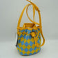 Yellow This everyday bucket bag features a distinctive argyle diamond pattern across its structured main body. Ideal for daily use, it is designed with a long, adjustable shoulder strap connected via metallic side rings. The spacious main storage compartment features an open-top design that closes with a functional drawstring woven through integrated metallic grommets along the upper rim, securing at the front with a knotted tie accent.